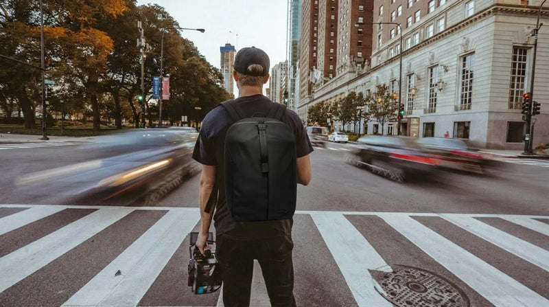 Man standing at city crosswalk wearing a black premium backpack with motion-blurred traffic