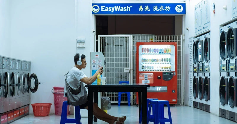 A man sitting with his shoulder bag in a late-night laundromat, flanked by rows of washers and dryers